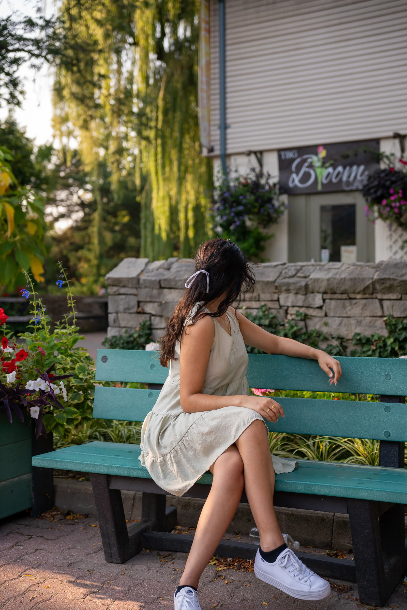 A woman sitting on a green bench in a blooming garden in a light summer dress at golden hour.